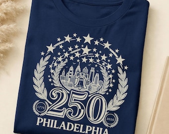 May include: A navy blue t-shirt featuring a white graphic celebrating Philadelphia's 250th anniversary. The design includes a cityscape, stars, laurel leaves, and the numbers "250" and the years "1776" and "2026".