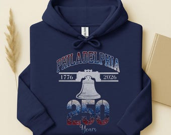 May include: A navy blue hoodie featuring a graphic design with the word "PHILADELPHIA" in red, white, and blue. The design also includes the Liberty Bell, the years "1776" and "2026", and the number "250".