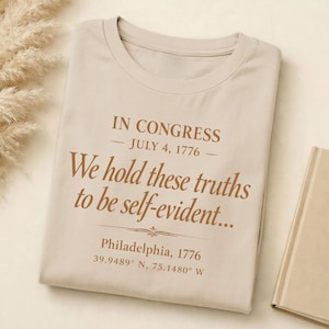 May include: Beige t-shirt with the text "IN CONGRESS - JULY 4, 1776 - We hold these truths to be self-evident..." in brown lettering. The shirt also includes "Philadelphia, 1776" and coordinates.