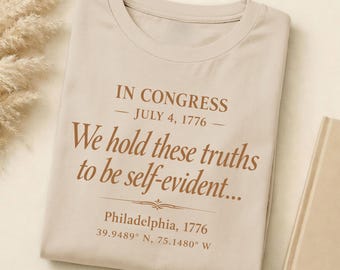 May include: Beige t-shirt with the text "IN CONGRESS - JULY 4, 1776 - We hold these truths to be self-evident..." in brown lettering. The shirt also includes "Philadelphia, 1776" and coordinates.