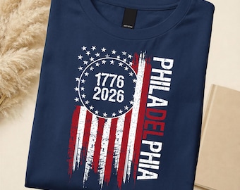 May include: Navy blue t-shirt with a distressed American flag design. The shirt features the text "1776 2026" inside a circle of stars. The word "PHILADELPHIA" is printed vertically in white and red.