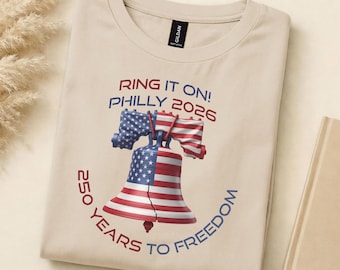 May include: Beige t-shirt with a graphic of the Liberty Bell in red, white, and blue, and the text "RING IT ON! PHILLY 2026" and "250 YEARS TO FREEDOM". The shirt is made of a soft material and is folded.