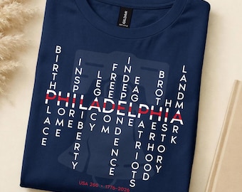 May include: Dark blue t-shirt featuring a white and red graphic. The word "PHILADELPHIA" is in red, surrounded by white words. The text "USA 250 - 1776-2026" is printed at the bottom of the shirt.