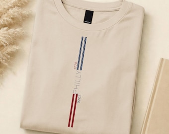 May include: A beige crew neck t-shirt, neatly folded. It has a vertical design with the word "PHILLY" and the numbers "1776" and "2026" in blue and red. The t-shirt is made of a soft material. The design is reminiscent of the American flag.
