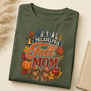 May include: An olive green t-shirt featuring a graphic print with the text "PHILADELPHIA Fall MOM". The design includes a city skyline, the Liberty Bell, a pumpkin, an apple, autumn leaves, and a plaid bow in shades of orange, red, and brown.