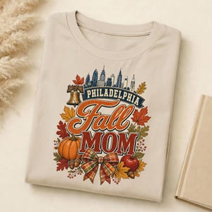 May include: A beige t-shirt featuring a graphic print of the Philadelphia skyline, a Liberty Bell, a pumpkin, an apple, acorns, and a plaid bow. The text reads Philadelphia Fall MOM in orange, red, and blue lettering surrounded by autumn leaves.
