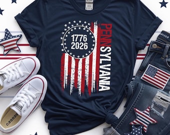 May include: Navy blue t-shirt with a distressed American flag design, the text "1776 2026" in a circle of stars, and the word "Pennsylvania". The shirt is styled with white trainers and blue jeans.