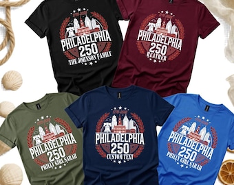 May include: Five short-sleeved crew neck t-shirts in black, maroon, olive green, navy blue, and royal blue. Each shirt features a white and red graphic of the Philadelphia skyline, the Liberty Bell, the text 1776-2026, PHILADELPHIA 250, and custom text.