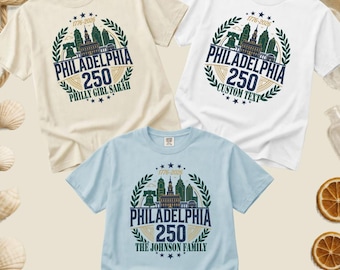 May include: Three short-sleeved t-shirts in cream, white, and light blue. Each features a circular graphic with a Philadelphia skyline, the Liberty Bell, and the text 1776-2026 PHILADELPHIA 250. The shirts include custom text at the bottom.