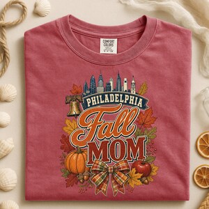 May include: A dusty rose crewneck t-shirt featuring a graphic of the Philadelphia skyline, a Liberty Bell, a pumpkin, an apple, and a plaid bow. The text reads Philadelphia Fall Mom. The shirt has a Comfort Colors brand label at the inner neckline.