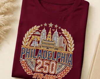 May include: A maroon t-shirt featuring a rhinestone graphic of the Philadelphia skyline, the Liberty Bell, and laurel wreaths. The design includes the text PHILADELPHIA, 250, and 1776-2026 in blue, gold, and clear crystals with five stars above.