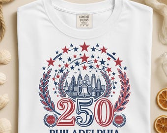 May include: White t-shirt with a circular design featuring the Philadelphia skyline, the Liberty Bell, and the number "250" in red and blue. The design includes stars and the text "PHILADELPHIA" below.