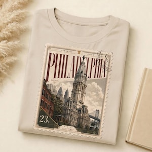 May include: A beige t-shirt featuring a vintage-style Philadelphia postage stamp design. The stamp showcases a detailed illustration of a historic building, the word "PHILADELPHIA" in bold letters, and the number "23".