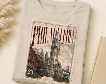 May include: A cream-coloured t-shirt with a vintage Philadelphia postage stamp design. The stamp features a detailed illustration of a historic building, the word "PHILADELPHIA", and the number "23".
