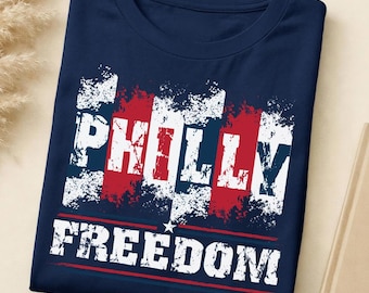 May include: A navy blue t-shirt featuring a distressed graphic print with vertical red and white stripes. The text "PHILLY" appears in large white block letters, followed by the word "FREEDOM" in white, separated by a small white star.