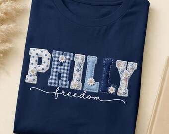 May include: A navy blue t-shirt featuring the word PHILLY in large, multi-patterned fabric letters with blue and white polka dots, gingham, floral prints, and solid light blue. Small white daisy appliques are attached to the letters. The word freedom is written below in white cursive script.