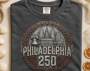 May include: A charcoal grey t-shirt featuring a circular graphic with a metallic silver Philadelphia skyline, the Liberty Bell, and the text Philadelphia 250 1776-2026. The design is framed by a bronze-toned laurel wreath and five stars at the top.