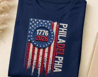 May include: A navy blue t-shirt featuring a textured, embroidered graphic of an American flag with a braided design. A circular blue emblem in the centre displays the numbers 1776 and 2026 in red and white. The word PHILADELPHIA is printed vertically.