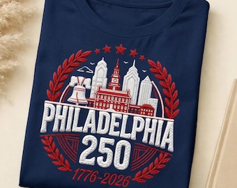 May include: A navy blue t-shirt with a white and red embroidered design. The design features the text "PHILADELPHIA 250" and the years "1776-2026", along with a depiction of the Philadelphia skyline, including the Liberty Bell.
