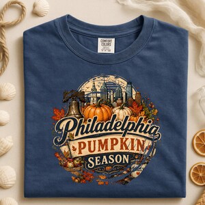 May include: A navy blue t-shirt featuring a graphic print of the Philadelphia skyline, the Liberty Bell, and pumpkins surrounded by autumn leaves. The text reads Philadelphia PUMPKIN SEASON. The shirt has a Comfort Colors brand label at the neck.