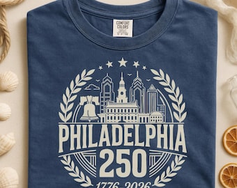 May include: A navy blue t-shirt featuring a white graphic of the Philadelphia skyline, the Liberty Bell, and the text "PHILADELPHIA 250 1776-2026". The shirt has a crew neck and short sleeves.