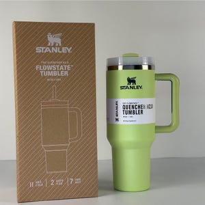 May include: A green Stanley Flowstate Quencher H2.0 tumbler with a handle and a lid, next to its cardboard box. The box features a line drawing of the tumbler and the Stanley logo. The tumbler holds 40 oz.