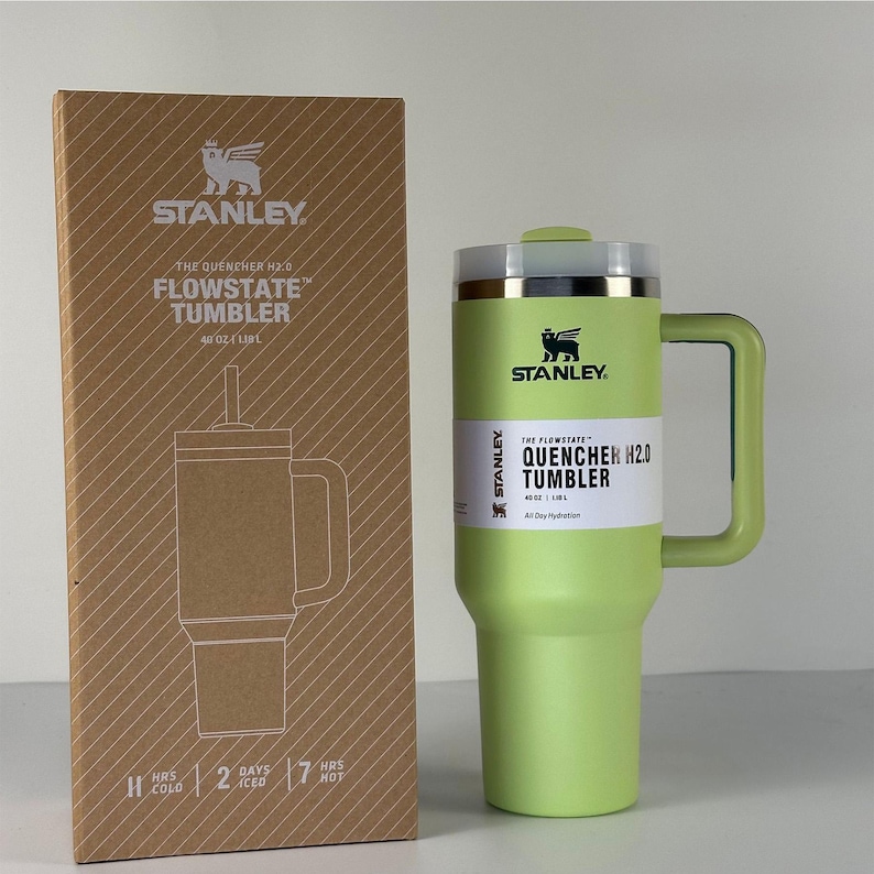 May include: A green Stanley Flowstate Quencher H2.0 tumbler with a handle and a lid, next to its cardboard box. The box features a line drawing of the tumbler and the Stanley logo. The tumbler holds 40 oz.