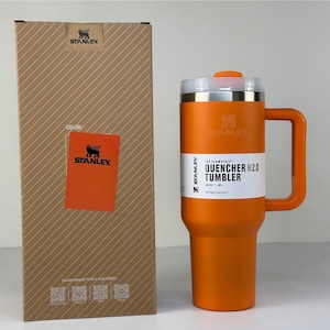 May include: An orange Stanley Quencher H2.0 tumbler with a handle, next to its cardboard box. The tumbler has a white label with the text "QUENCHER H2.0 TUMBLER". The box has the Stanley logo and the word "COLOR" on it.