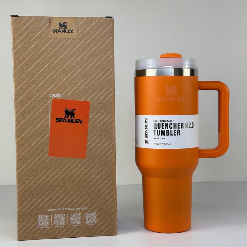 May include: An orange Stanley Quencher H2.0 tumbler with a handle, next to its cardboard box. The tumbler has a white label with the text "QUENCHER H2.0 TUMBLER". The box has the Stanley logo and the word "COLOR" on it.