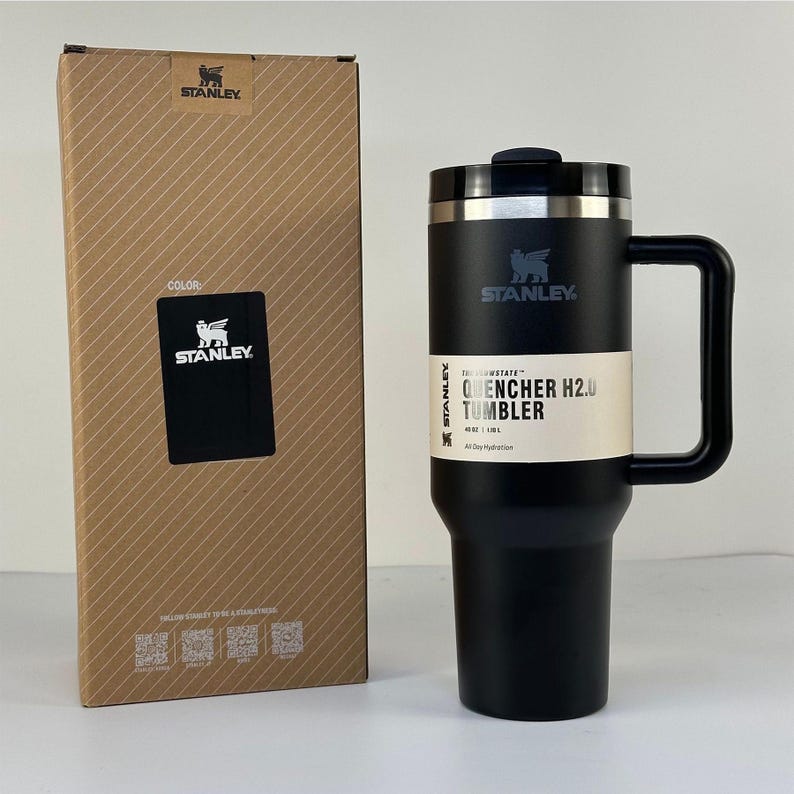 May include: A black Stanley Quencher H2.0 tumbler with a handle and a lid, next to its cardboard box. The tumbler has the Stanley logo and the text "QUENCHER H2.0 TUMBLER". The box has the Stanley logo and the word "COLOR".