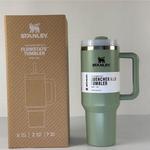 May include: A green Stanley Flowstate Quencher H2.0 tumbler with a handle and a straw, next to its cardboard box. The box features a drawing of the tumbler and the Stanley logo. The tumbler holds 48 oz.