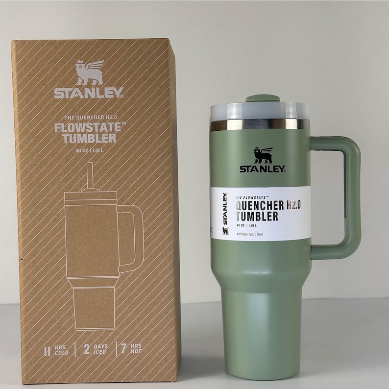 May include: A green Stanley Flowstate Quencher H2.0 tumbler with a handle and a straw, next to its cardboard box. The box features a drawing of the tumbler and the Stanley logo. The tumbler holds 48 oz.