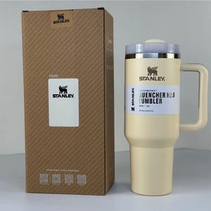 May include: A beige Stanley Quencher H2.0 tumbler with a handle, next to its matching cardboard box. The tumbler has a silver rim and a white label that reads "QUENCHER H2.0 TUMBLER" and "All Day Hydration". The box has the Stanley logo and a color label.