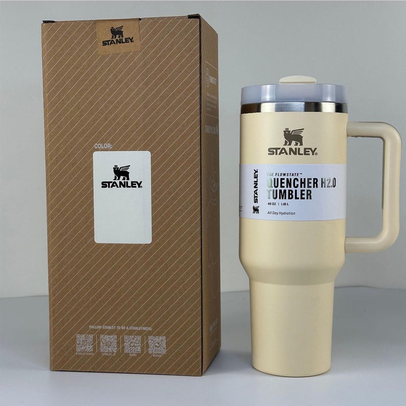 May include: A beige Stanley Quencher H2.0 tumbler with a handle, next to its matching cardboard box. The tumbler has a silver rim and a white label that reads "QUENCHER H2.0 TUMBLER" and "All Day Hydration". The box has the Stanley logo and a color label.