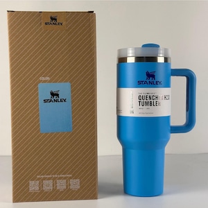 May include: A blue Stanley Quencher H2.0 tumbler with a handle and a lid, next to its cardboard box. The tumbler has the Stanley logo and the text "QUENCHER H2.0 TUMBLER". The box has the Stanley logo and a color swatch.