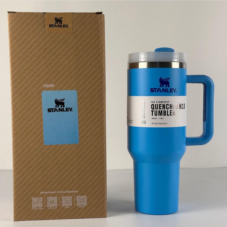 May include: A blue Stanley Quencher H2.0 tumbler with a handle and a lid, next to its cardboard box. The tumbler has the Stanley logo and the text "QUENCHER H2.0 TUMBLER". The box has the Stanley logo and a color swatch.
