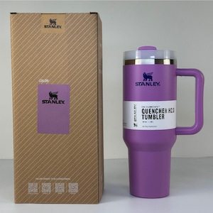 May include: A purple Stanley Quencher H2.0 tumbler with a handle and a lid, next to its cardboard box. The box has a color label that says "COLOR:" and "STANLEY". The tumbler has the text "THE FLOWSTATE QUENCHER H2.0 TUMBLER" on it.