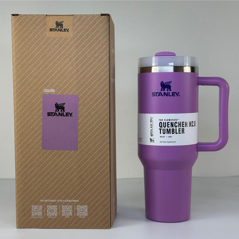 May include: A purple Stanley Quencher H2.0 tumbler with a handle and a lid, next to its cardboard box. The box has a color label that says "COLOR:" and "STANLEY". The tumbler has the text "THE FLOWSTATE QUENCHER H2.0 TUMBLER" on it.
