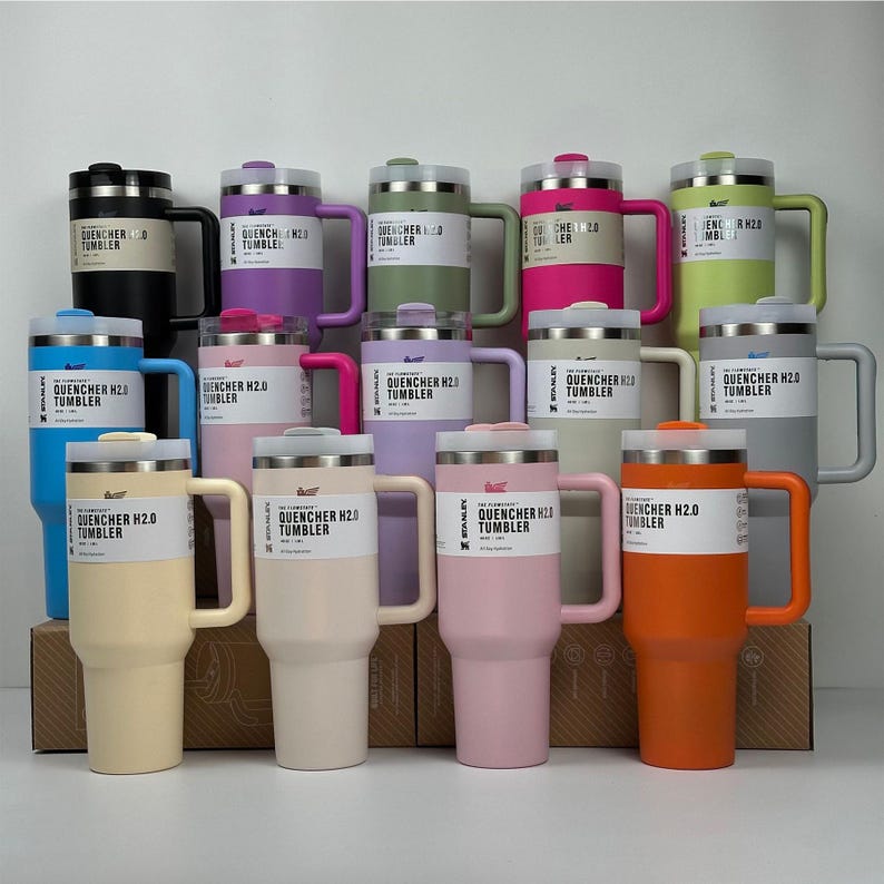 May include: A collection of insulated tumblers in various colors, including black, purple, green, pink, blue, and orange. Each tumbler has a handle and a lid, with the text "QUENCHER H2.0 TUMBLER" printed on a label. The tumblers are arranged on a white surface.