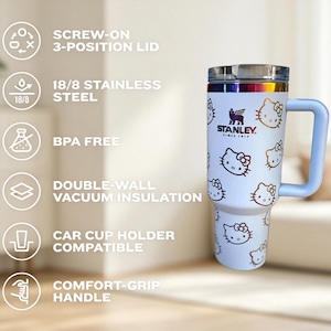 May include: A white Stanley travel mug featuring Hello Kitty graphics. The mug has a light blue handle and a screw-on lid. The text on the mug reads "STANLEY SINCE 1913". The mug is made of stainless steel and is car cup holder compatible.