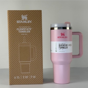 May include: A pink Stanley Quencher H2.0 Flowstate tumbler with a handle, 40 oz (1.18L) capacity. The tumbler has a white and silver lid and a white label with the product name. The box next to it has the Stanley logo and product information.