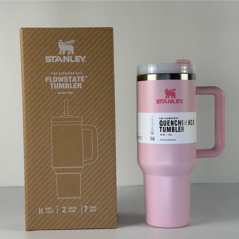 May include: A pink Stanley Quencher H2.0 Flowstate tumbler with a handle, 40 oz (1.18L) capacity. The tumbler has a white and silver lid and a white label with the product name. The box next to it has the Stanley logo and product information.
