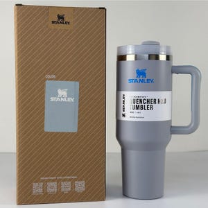 May include: A gray Stanley Quencher H2.0 tumbler with a handle, next to its cardboard box. The tumbler has a silver rim and the Stanley logo. The box is brown with diagonal lines and the Stanley logo.