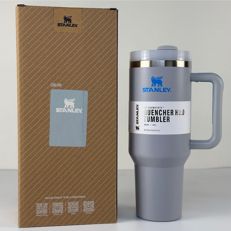 May include: A gray Stanley Quencher H2.0 tumbler with a handle, next to its cardboard box. The tumbler has a silver rim and the Stanley logo. The box is brown with diagonal lines and the Stanley logo.