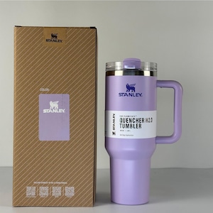 May include: A lavender Stanley Quencher H2.0 tumbler with a handle and a clear lid, next to a brown cardboard box. The tumbler has the Stanley logo and the text "QUENCHER H2.0 TUMBLER". The box has the Stanley logo and the word "COLOR".