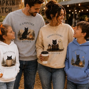 May include: A family of four wearing matching sweatshirts and hoodies featuring black cat illustrations. The white hoodie says Hot Cocoa Expert, the grey sweatshirt says Campfire Keeper, the beige sweatshirt says Ghost Story Teller, and the blue hoodie says Marshmallow Roaster.