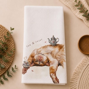 Puede incluir: Un paño de cocina blanco con tejido de gofre que presenta una ilustración en acuarela de un gato atigrado naranja tumbado boca arriba. El gato aparece bajo una tetera floral y junto a una pequeña taza de té. El texto "tea & reset" está impreso.