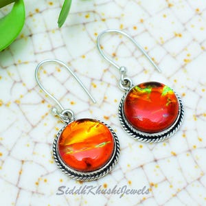 May include: A pair of drop earrings featuring round, vibrant orange dichroic glass cabochons set in twisted silver-tone metal frames. The earrings hang from simple silver-tone hooks. The text SiddhKhushiJewels is visible at the bottom of the image.