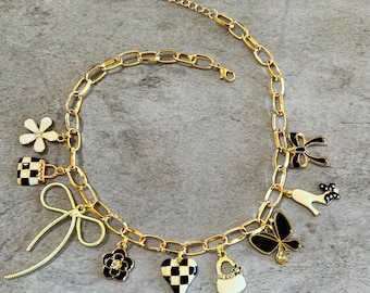 May include: Gold-coloured chain necklace with assorted charms, including a flower, bow, heart, purse, and butterfly. The charms feature black and white chequered patterns and enamel details. The necklace has a lobster clasp closure.