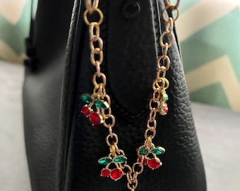 May include: A gold chain bag charm with red and green cherry charms. The cherries are made of rhinestones.